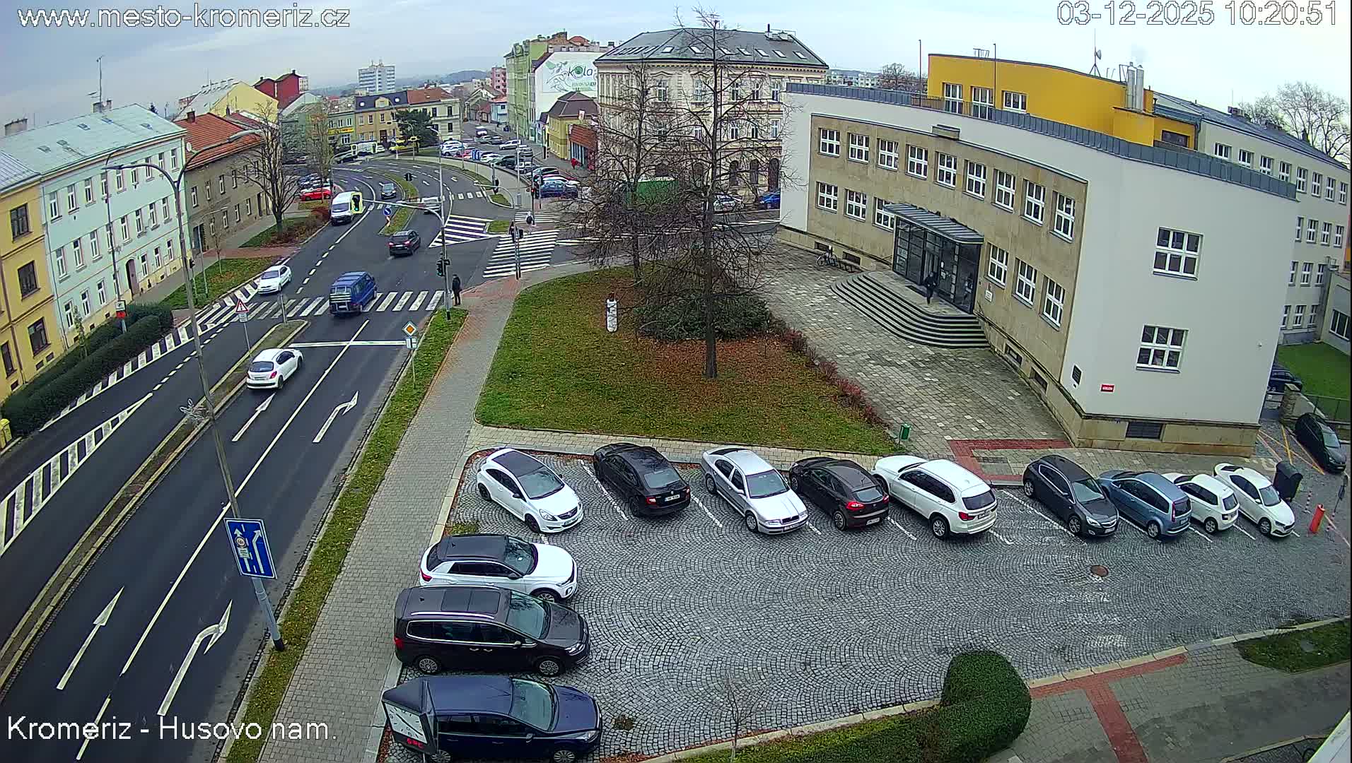 An overhead view captures a cloudy urban scene featuring a multi-lane street with ongoing traffic, several parked cars in a cobblestone lot, and various buildings under an overcast sky.