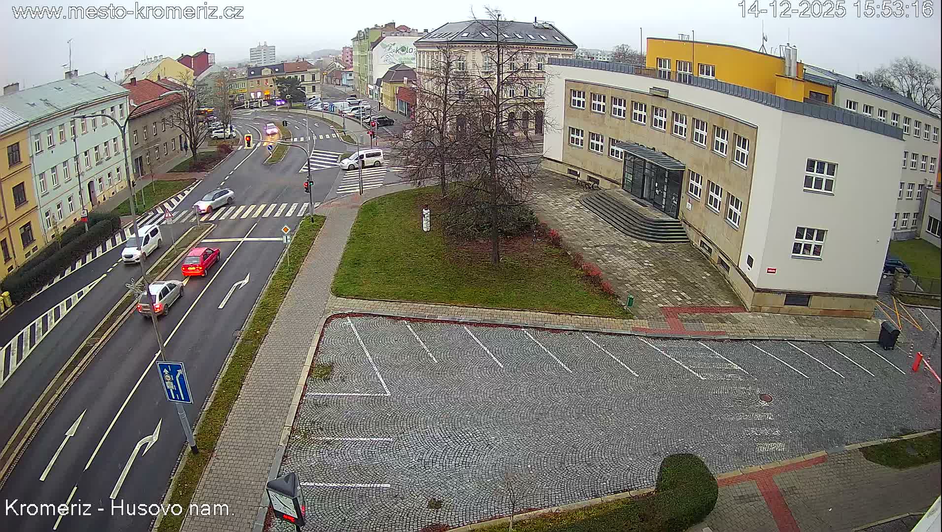 An overhead view captures a cloudy urban scene featuring a multi-lane street with ongoing traffic, several parked cars in a cobblestone lot, and various buildings under an overcast sky.