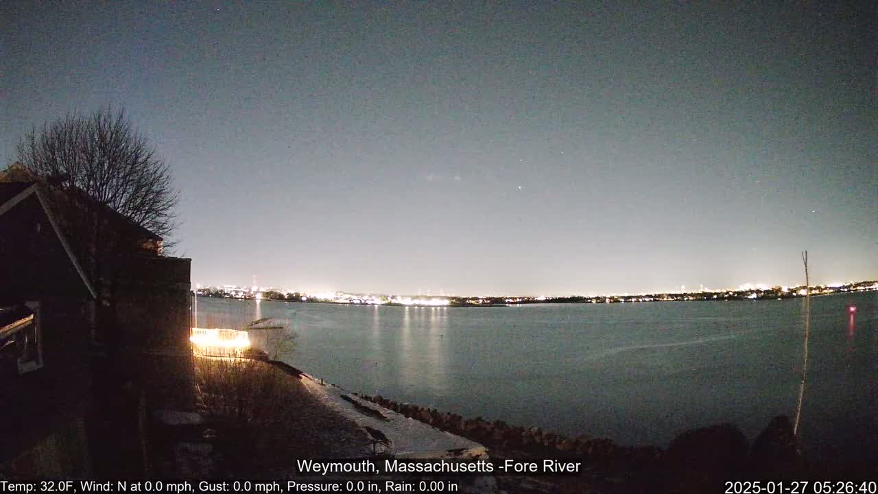 A nighttime view of a calm river with city lights on the opposite shore, seen from a snow-dusted shoreline near a dark house under a clear sky.