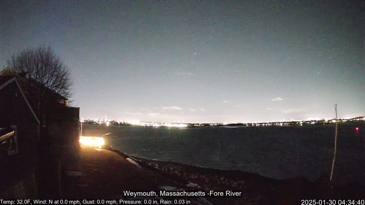 A nighttime view of a dark body of water with city lights on the horizon under a clear, dark sky, partially illuminated by a nearby house.