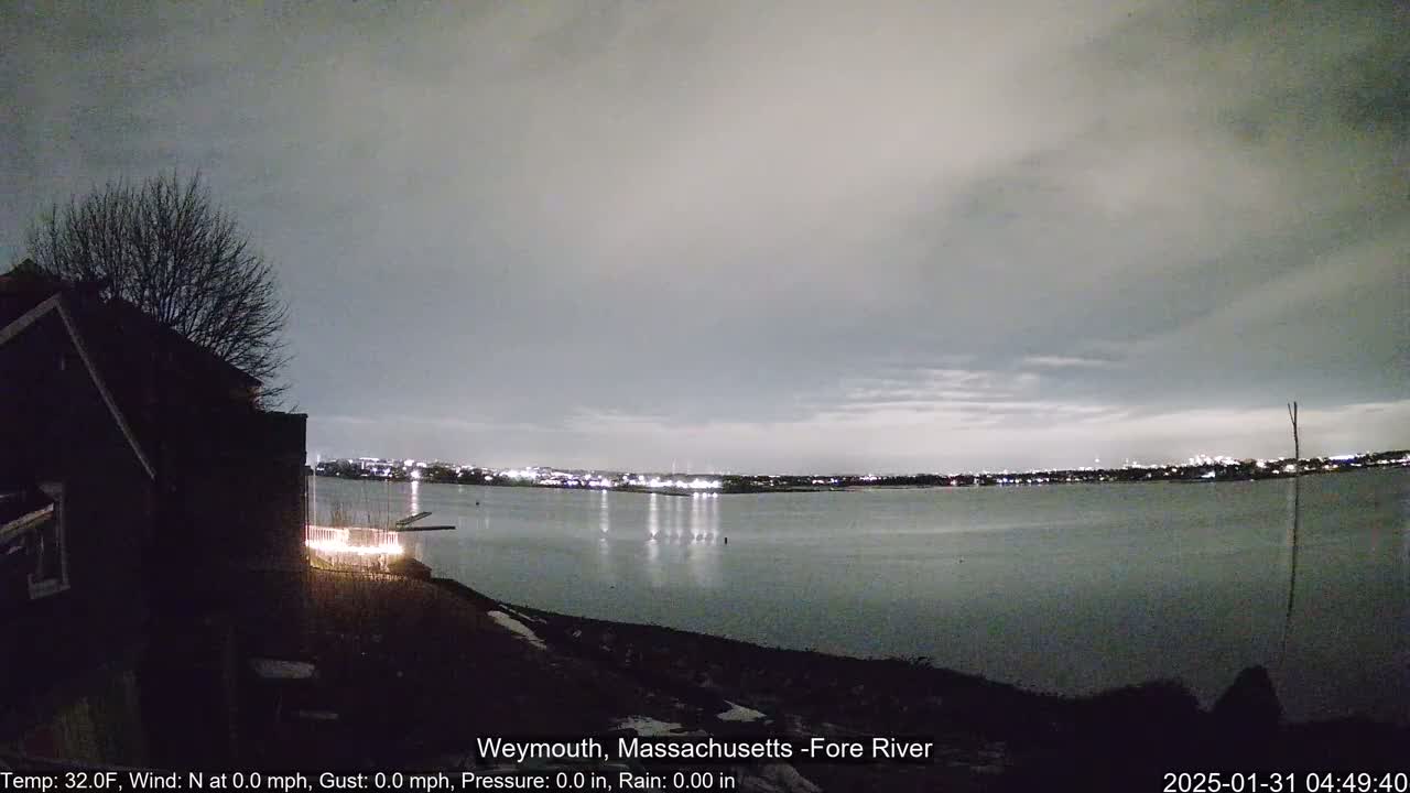 A nighttime view of a calm river, reflecting city lights across the water under an overcast sky.