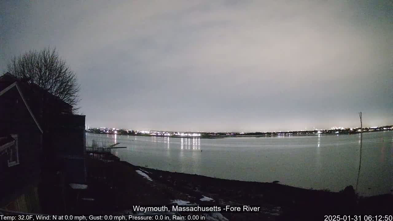 A nighttime view of a calm river reflecting city lights under a gray sky, with a dark house and tree in the foreground.