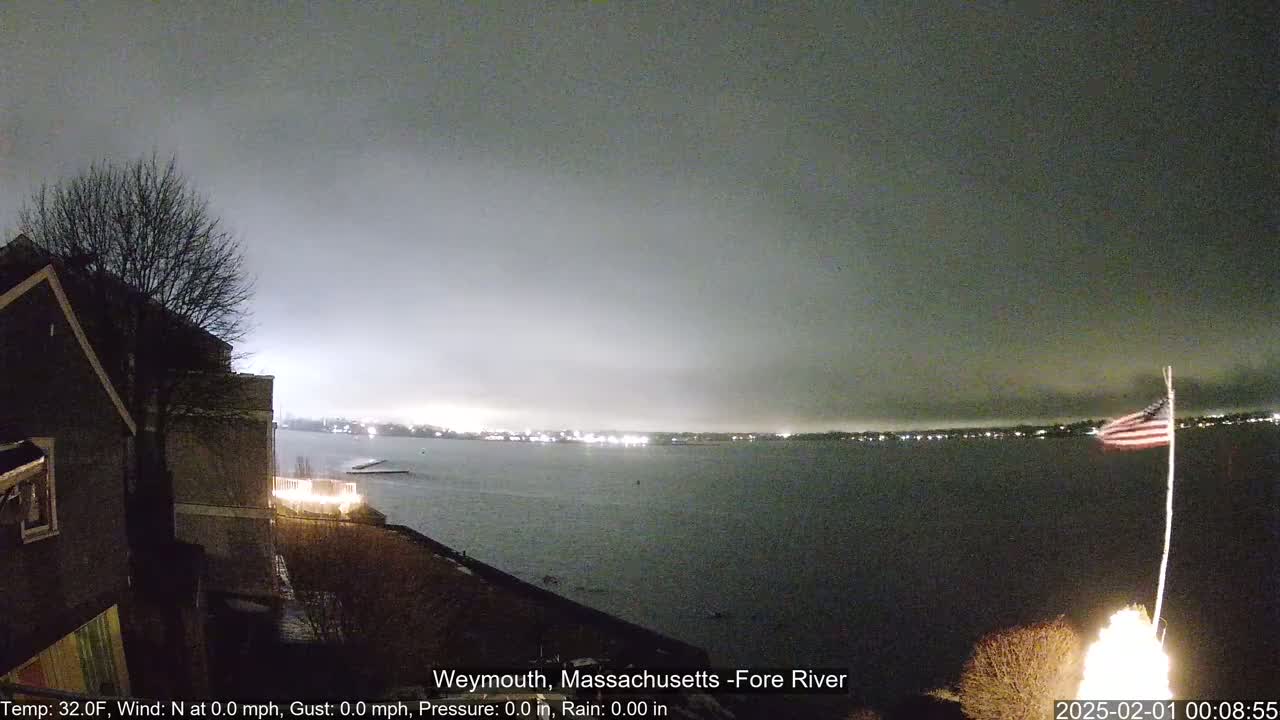 A nighttime view of a calm body of water under a cloudy sky, with a distant shoreline of lights and an American flag waving near a bonfire in the foreground.