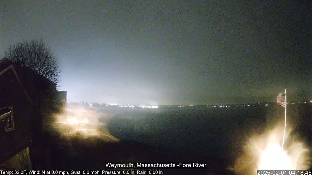 A nighttime view of a dark body of water with fireworks exploding over it, under a cloudy sky.