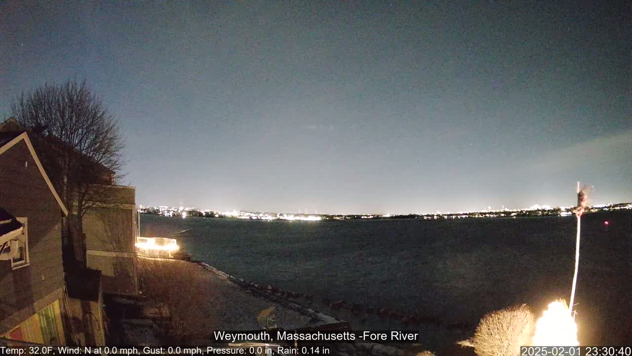 A nighttime view of a calm body of water with city lights on the far shore, under a dark sky, seen from a waterfront location near a house and bare trees.