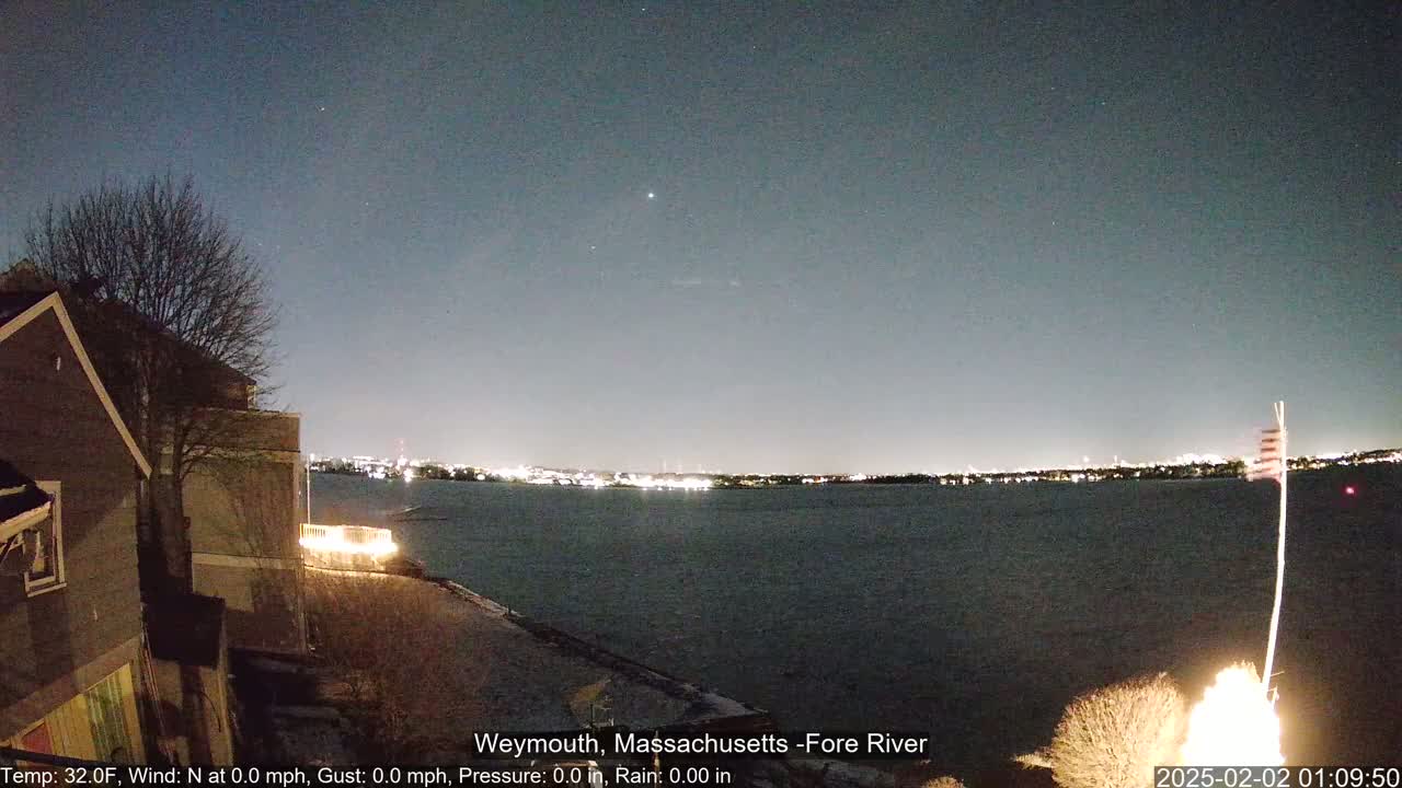 A nighttime view of a calm body of water with a city skyline in the distance, under a clear, dark sky with a few visible stars, from a vantage point near a house.