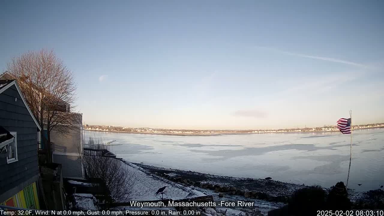 A partially frozen river is visible from a snowy shoreline, with a house and a bare tree in the foreground under a clear sky.
