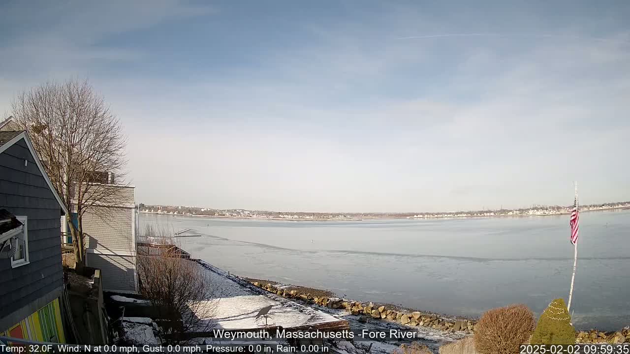 A partially frozen river is visible from a snowy shoreline, with houses and a bare tree in the foreground under a mostly clear sky.
