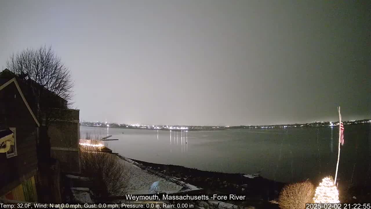A nighttime view of a calm river with a lightly snow-covered shoreline, showing a house and a lit-up Christmas tree in the foreground under a gray sky.