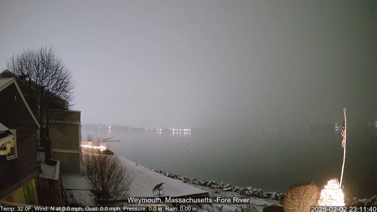 A snow-covered shoreline at night overlooks a calm, foggy river with distant lights.