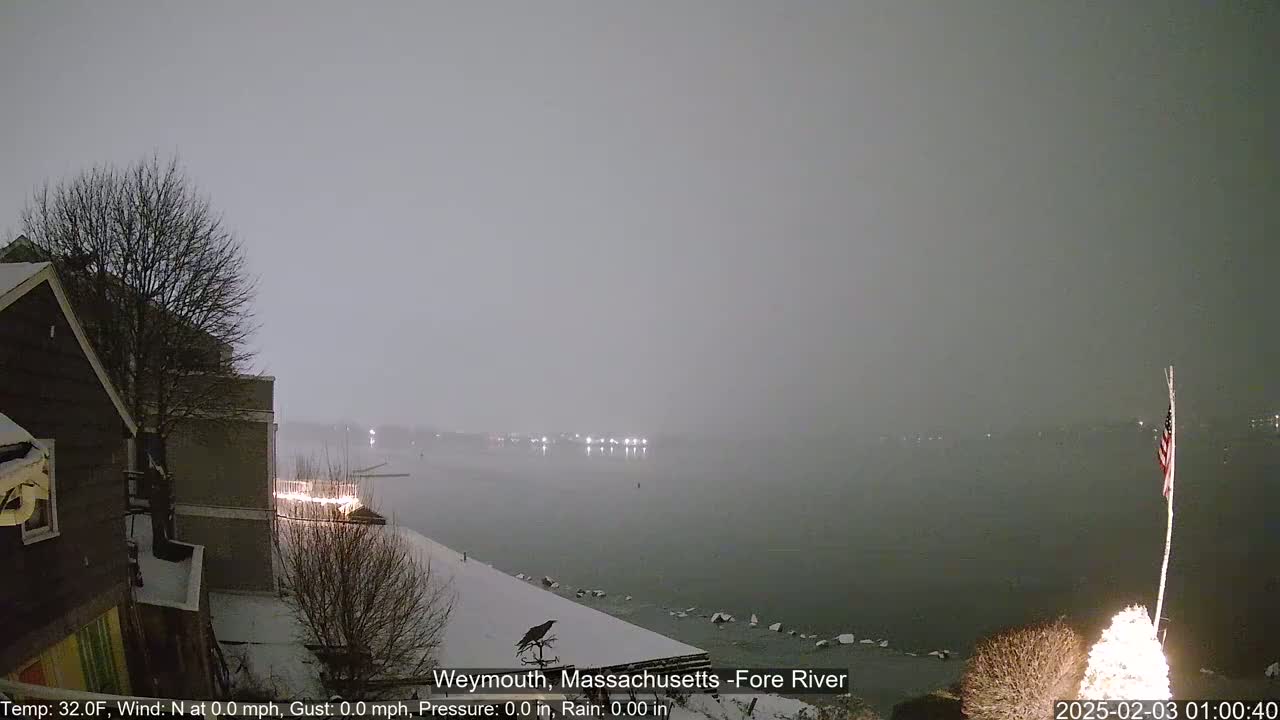 A snow-covered waterfront scene at night shows a calm body of water under a hazy sky, with buildings and a partially visible American flag in the foreground.