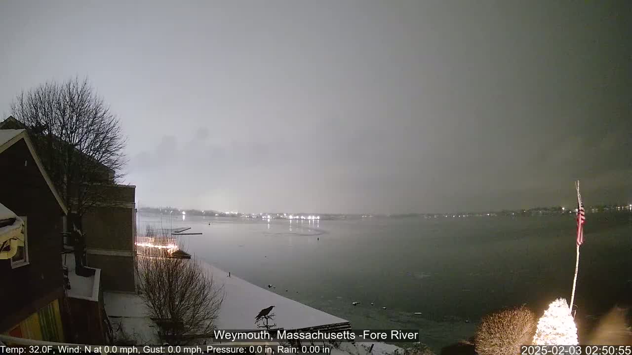 A nighttime view of a snow-covered shoreline overlooking a calm, dark river under a gray sky.