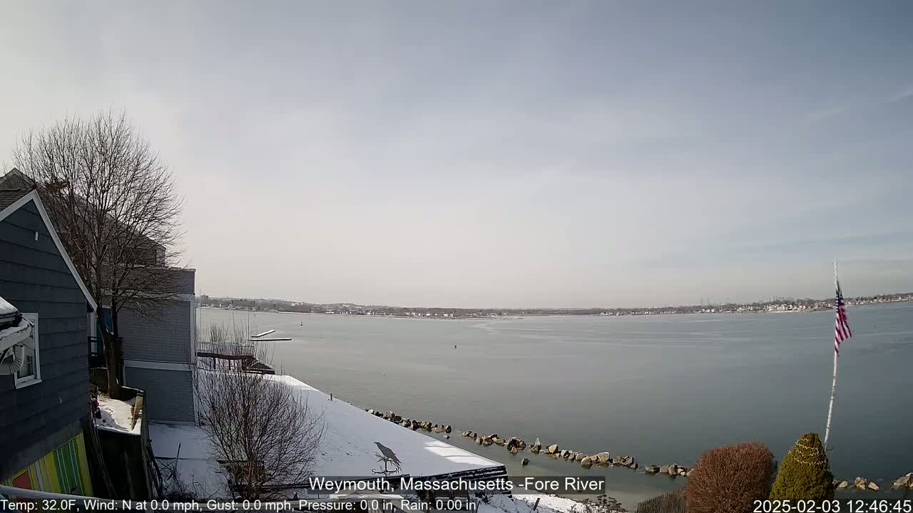 A snow-covered waterfront scene shows a calm body of water with a distant shoreline under a clear, light-grey sky.