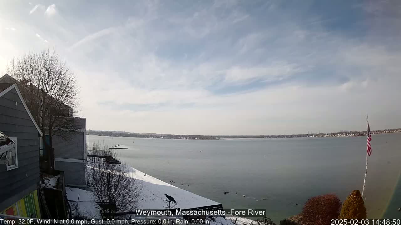 A gray house partially covered in snow overlooks a calm, gray body of water under a partly cloudy sky.
