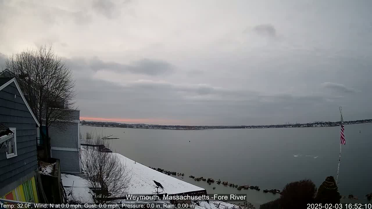 A snow-covered waterfront scene shows a calm, gray body of water under an overcast sky, with a gray house and a bare tree partially visible in the foreground.