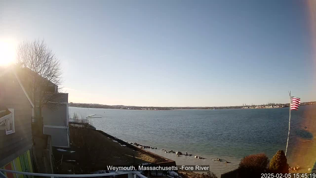 A sunny day reveals a wide body of water, with a distant shoreline and a small American flag on a pole in the foreground, near a house and sparsely vegetated land.