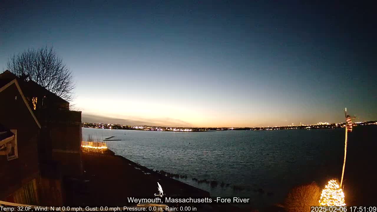 A dark house with lights on overlooks a calm body of water at twilight under a clear sky.