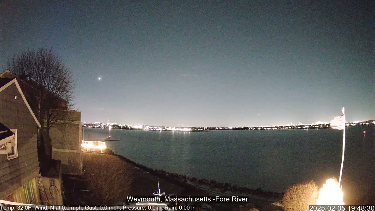 A nighttime view of a calm body of water with city lights in the distance, under a clear, dark sky, with a house and a bare tree in the foreground.