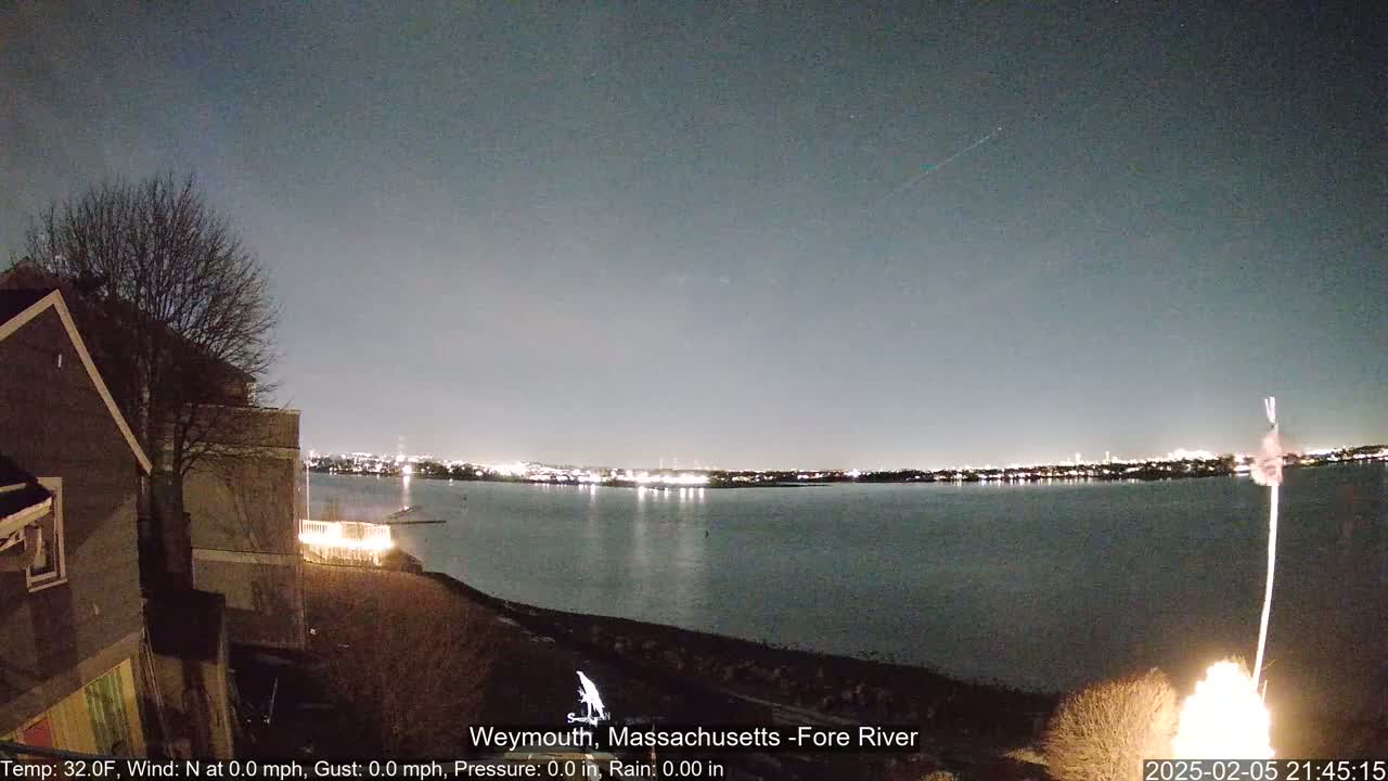 A nighttime view shows a calm body of water with city lights across the shore, a dark tree, a house, and a small fire in the foreground under a clear, dark sky with a meteor visible.