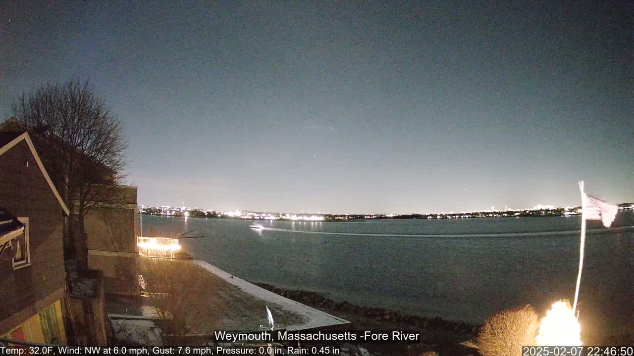 A nighttime view of a calm body of water with a city skyline in the distance, under a dark, clear sky, with a partially lit house and a flagpole in the foreground.