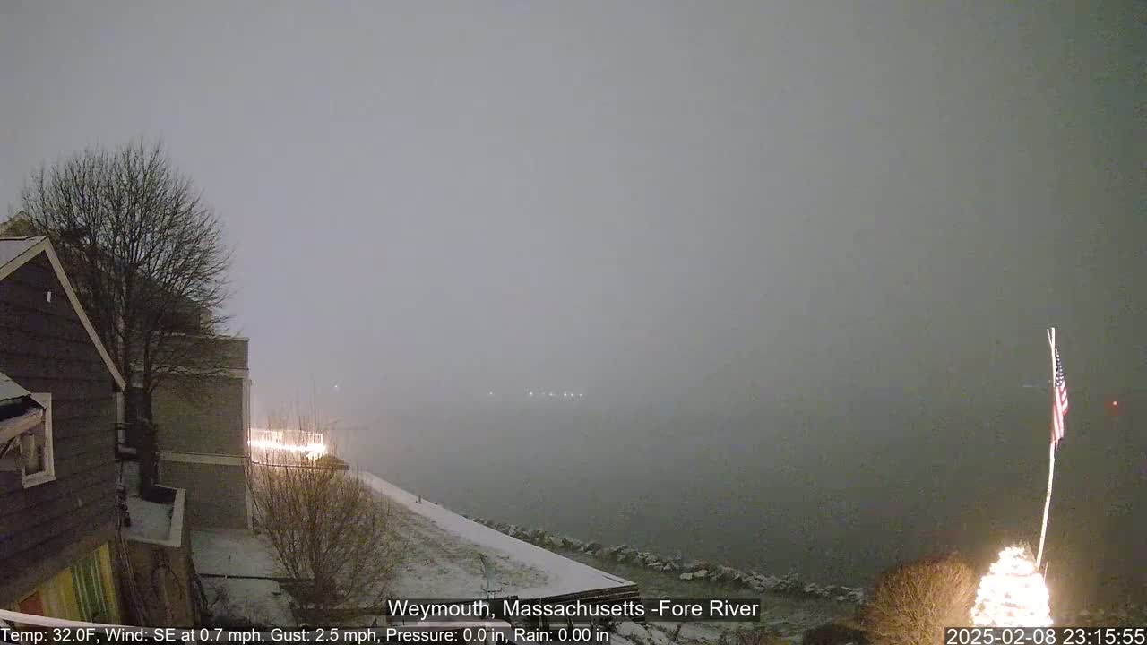 A snow-covered waterfront scene at night shows a partly obscured view of a body of water under a heavy, gray sky.