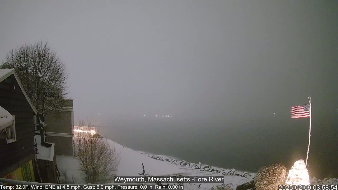A snow-covered waterfront scene shows a partially obscured view of a body of water under a heavy, gray, snowy sky.