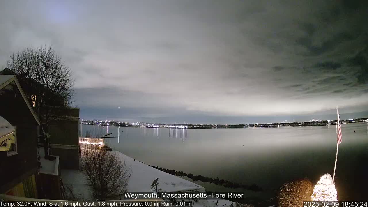 A snow-covered waterfront scene at night shows a calm body of water reflecting city lights under a cloudy sky.