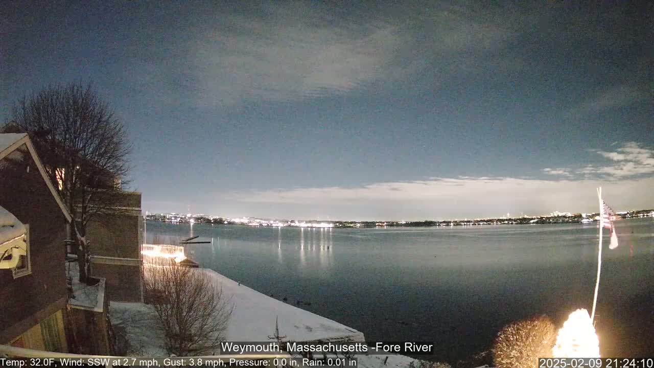 A nighttime view of a snow-covered waterfront, showing a calm river reflecting city lights under a partly cloudy sky.
