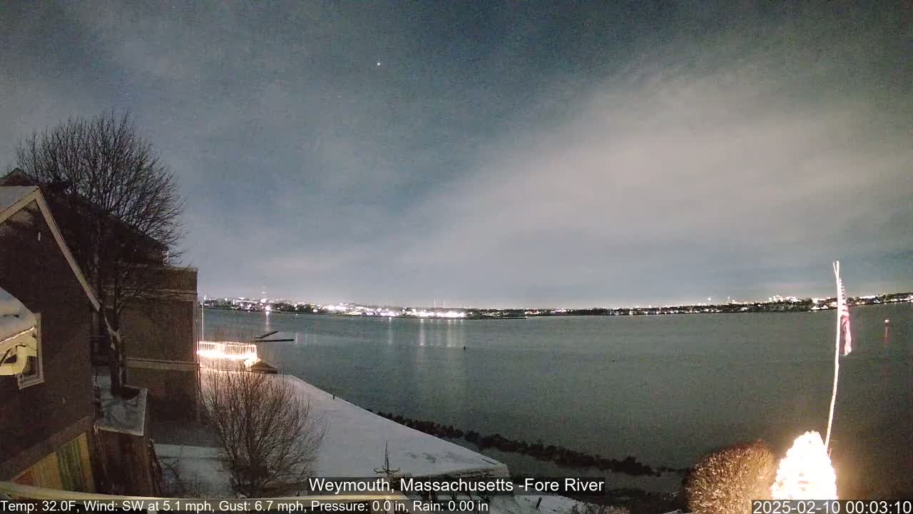 A nighttime view of a snow-covered waterfront with a calm river reflecting city lights under a cloudy sky.
