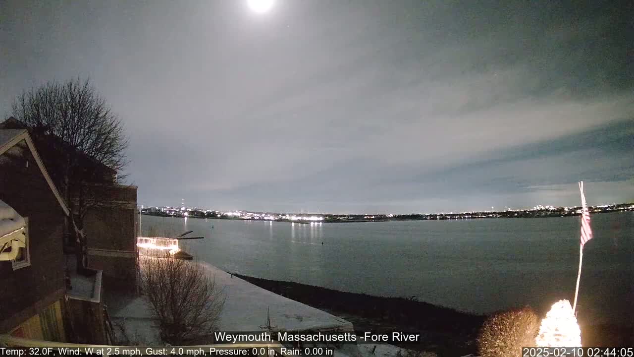 A nighttime view of a snow-covered waterfront, showing a body of water with city lights across it under a mostly cloudy sky illuminated by a bright moon.