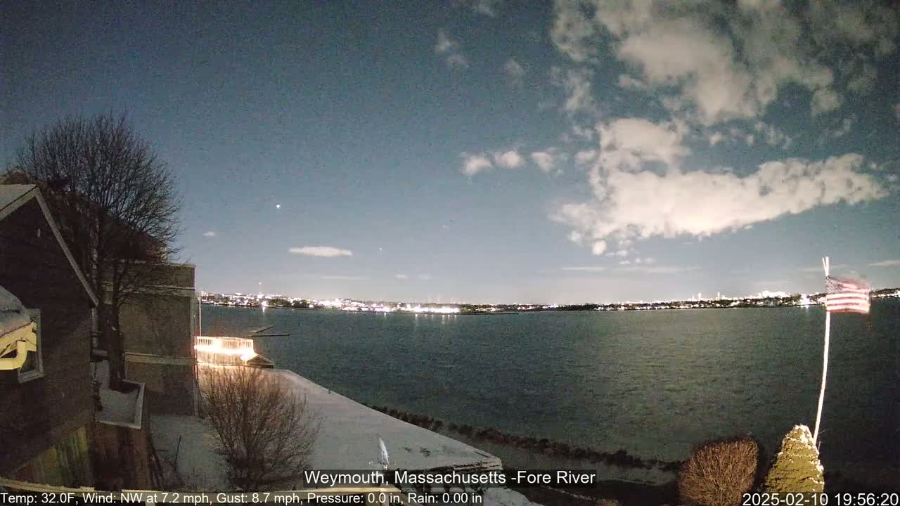A nighttime view of a snow-covered waterfront shows a calm body of water with city lights across it under a partly cloudy sky.