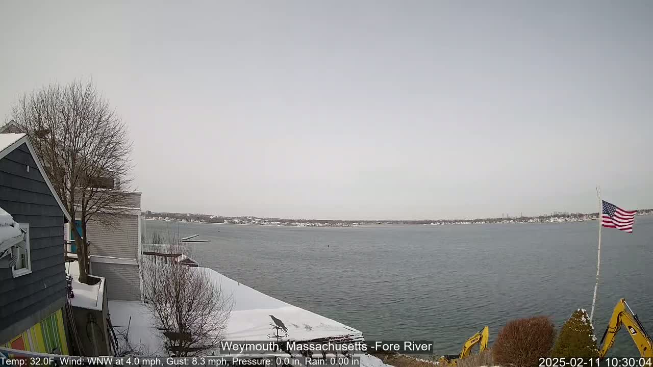 A snow-covered waterfront scene shows a body of water under a cloudy sky, with houses and construction equipment visible in the foreground.