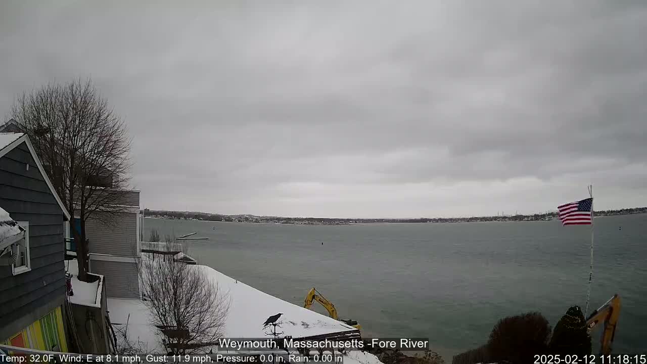 A snow-covered waterfront scene shows a calm body of water under an overcast sky, with houses, a construction vehicle, and an American flag visible.