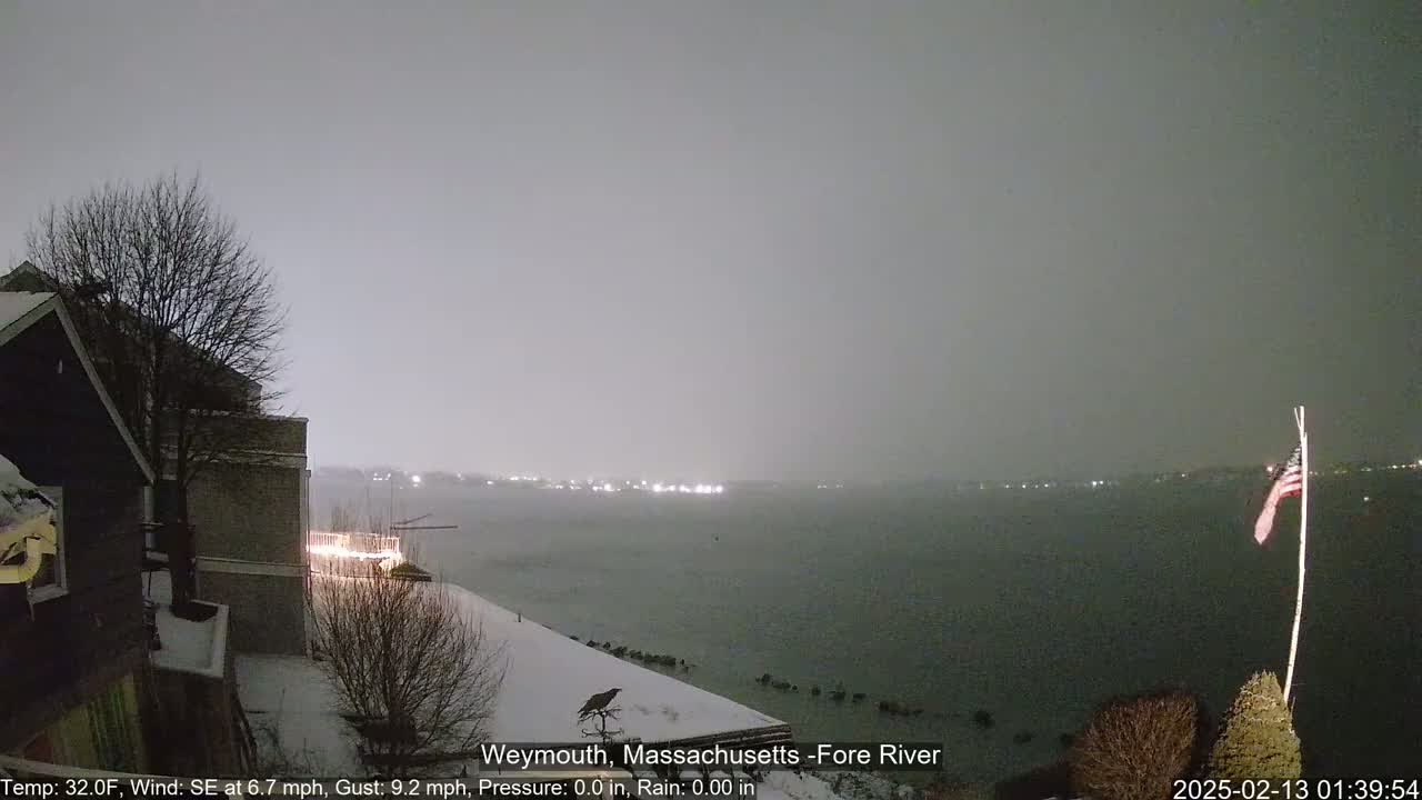 A snow-covered waterfront scene at night shows a dark building, a bare tree, and a partially visible American flag, with a calm, dark body of water under a gray, overcast sky.