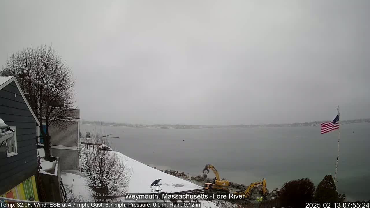 A snowy waterfront scene shows construction equipment working near a body of water under an overcast sky.