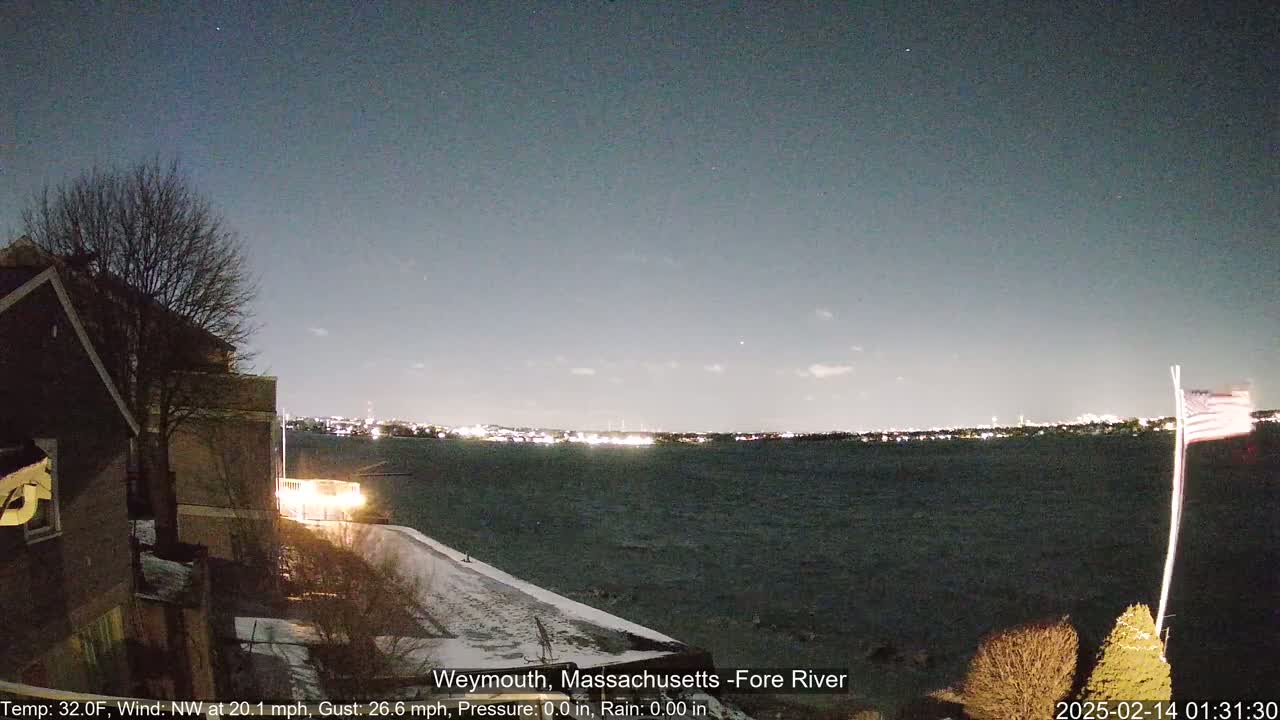 A nighttime view of a body of water with a city skyline in the distance, under a mostly clear sky, with snow on the ground in the foreground and a partially lit building to the left.