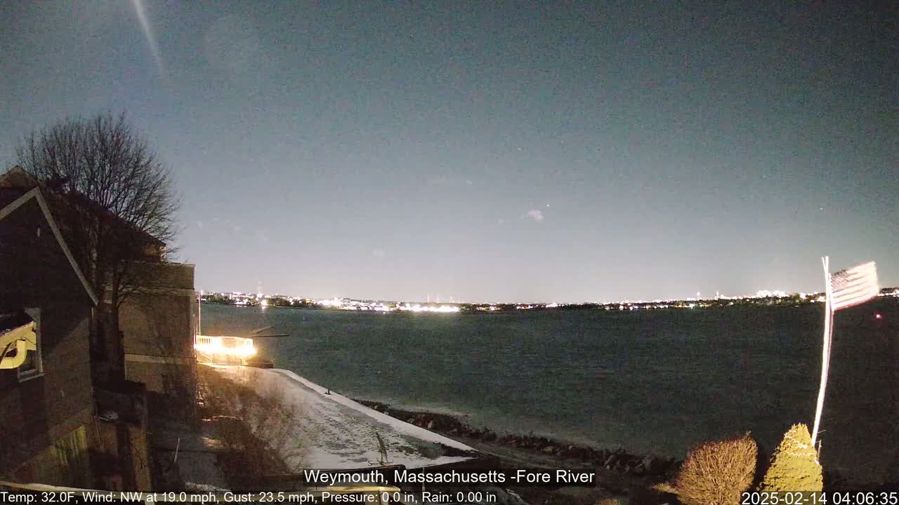 A nighttime view of a snow-dusted shoreline and a calm body of water, with city lights visible across the water under a clear, dark sky.