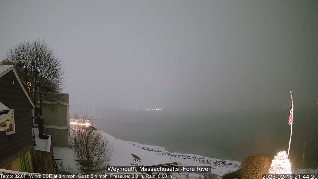 A snow-covered waterfront scene shows a partly visible house, bare trees, and a snow-covered shoreline extending to a foggy river under a gray sky during a light snowfall.