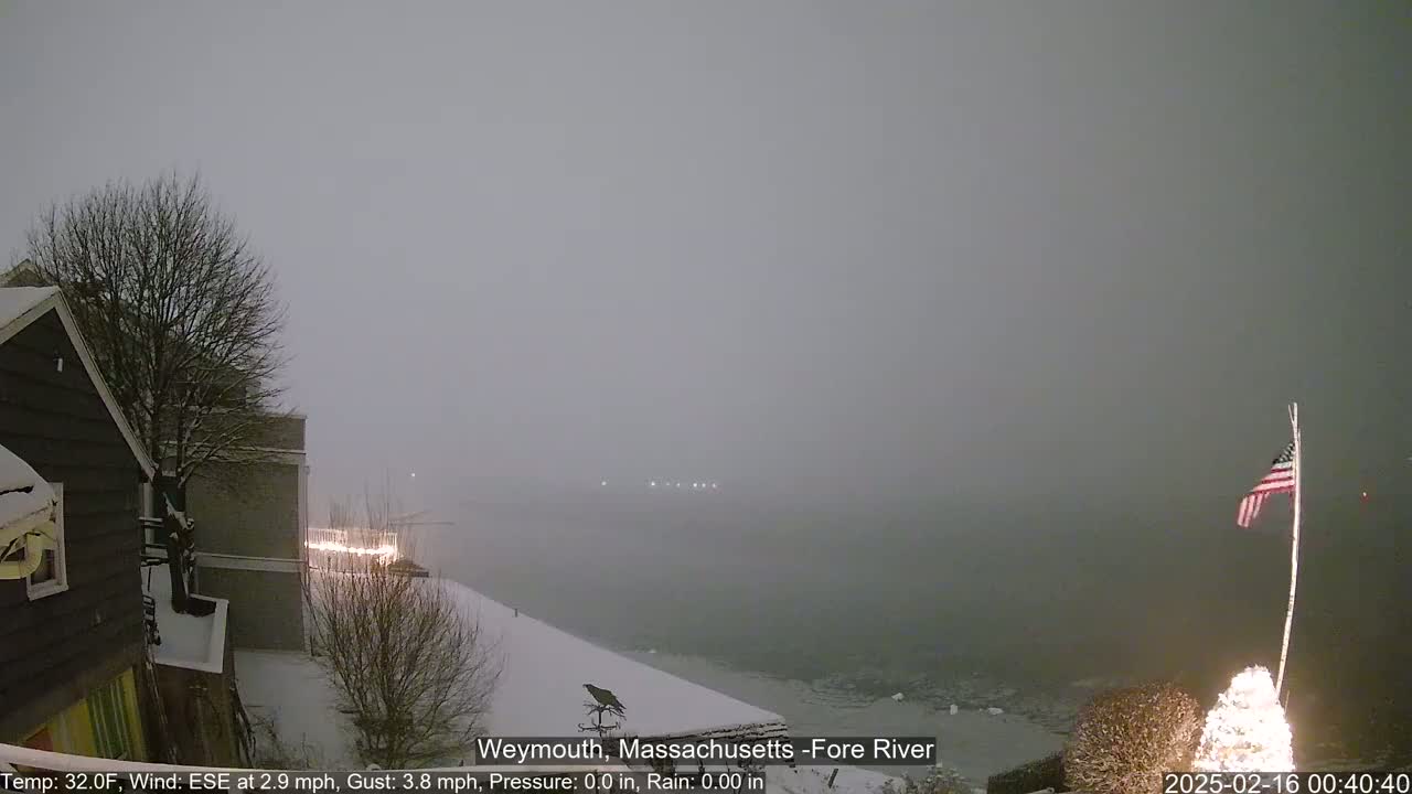 A snow-covered waterfront scene shows a partly visible building, a bare tree, and a river under a heavy, gray sky.