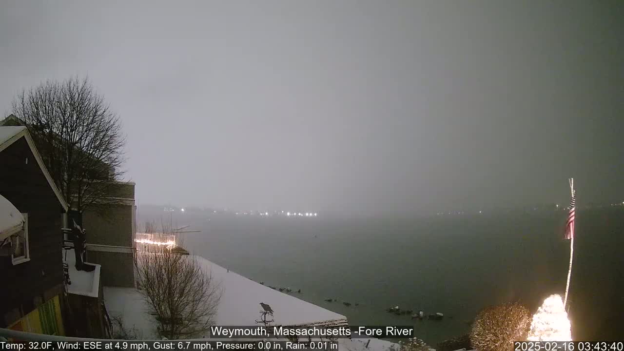 A snow-covered waterfront scene at night shows a body of water under a heavy, gray sky, with a snow-covered building and a lit-up Christmas tree in the foreground.
