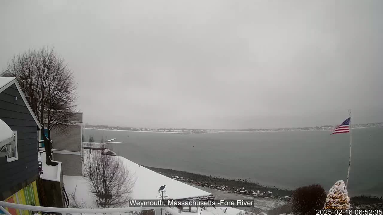 A snow-covered residential area overlooks a calm, grey river under a cloudy sky.