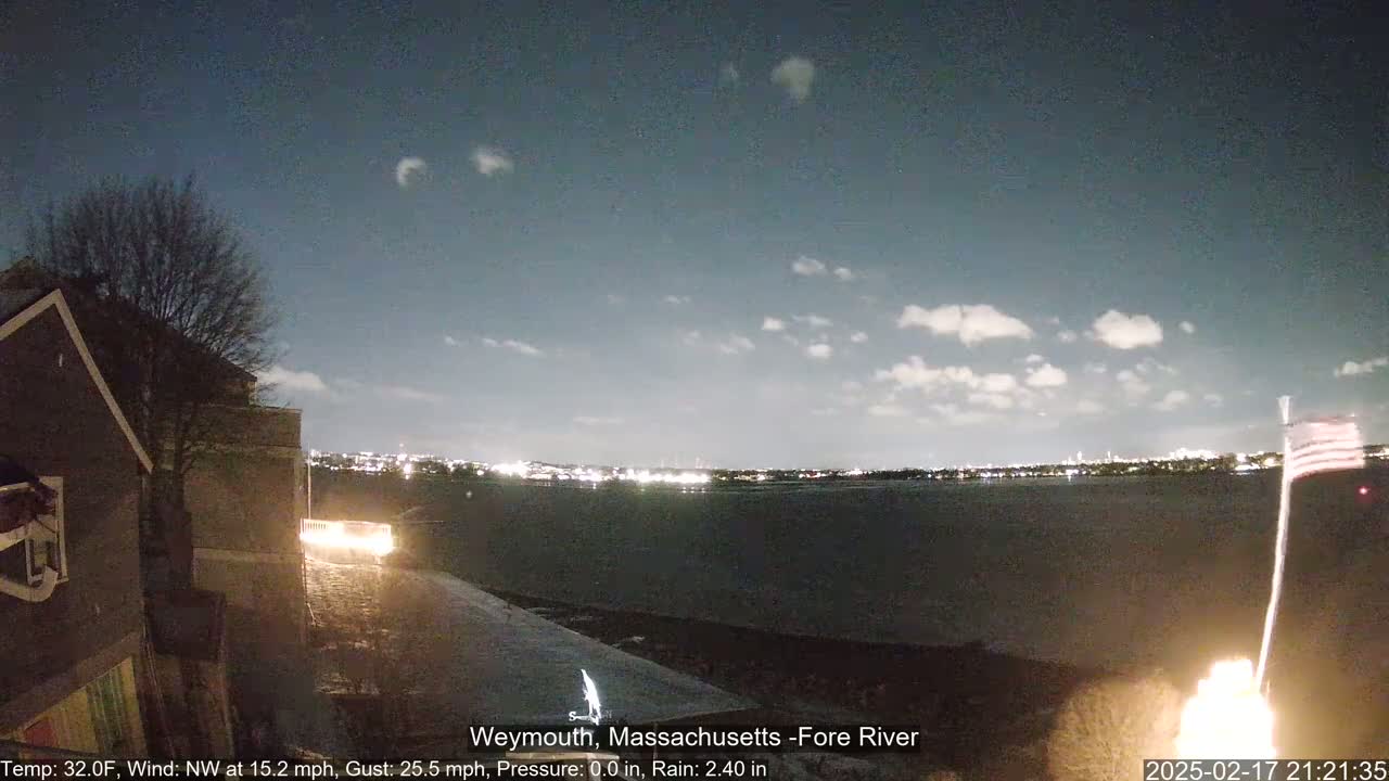 A nighttime view of a body of water with city lights across it, under a partly cloudy sky, from a vantage point near a house with a small American flag visible.
