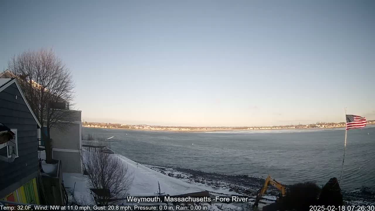 A partly frozen river is seen from a snowy shoreline with a house and an American flag in the foreground under a clear, bright sky.