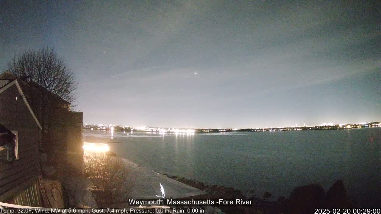 A nighttime view of a calm river with city lights on the far shore, seen from a snow-covered embankment near a house under a clear, dark sky.