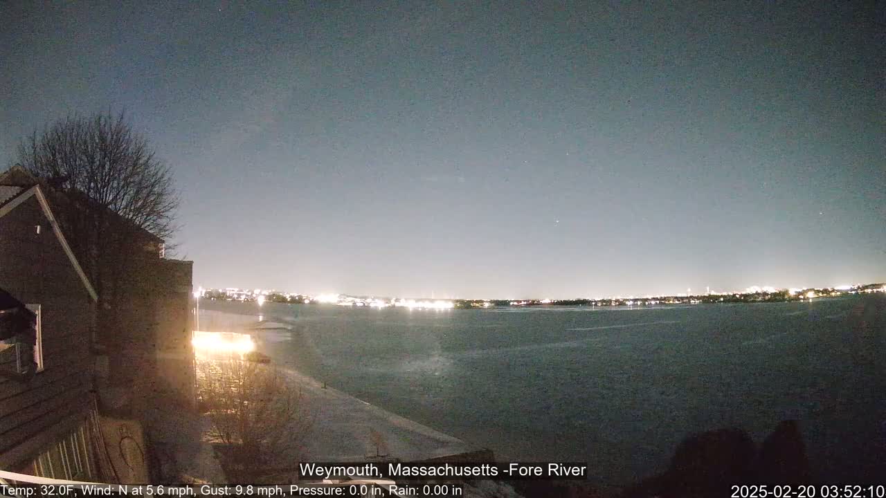 A nighttime view of a dark body of water with city lights across it, seen from a snow-covered shoreline beside a dark building under a clear, dark sky.
