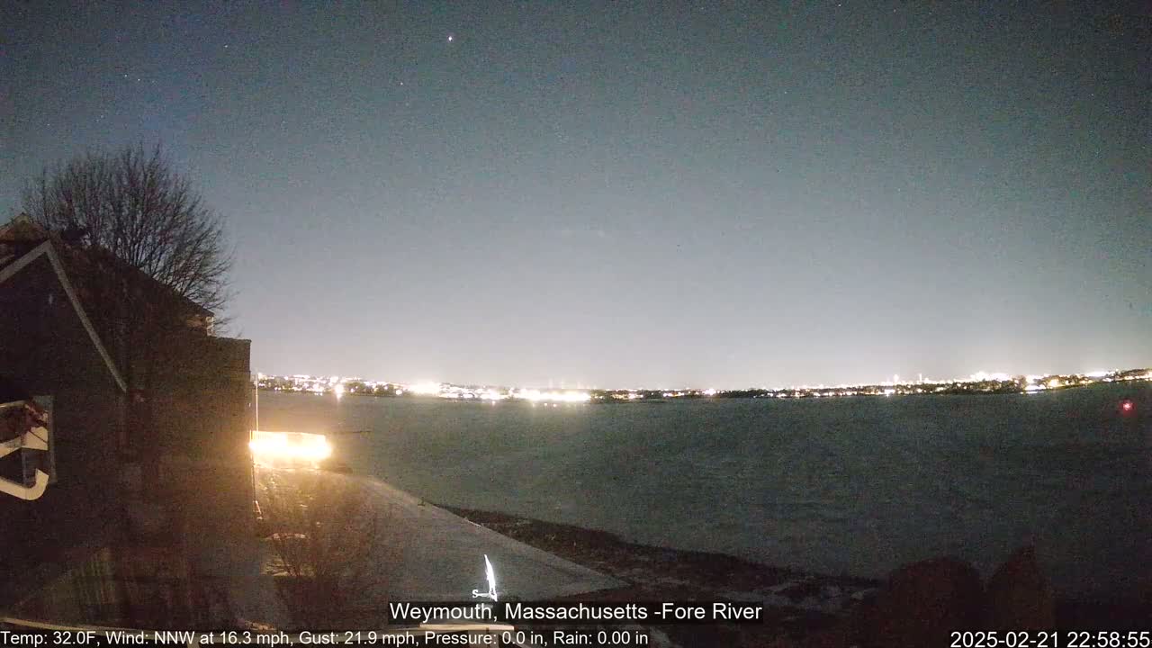 A nighttime view of a calm body of water with a distant city skyline illuminated under a clear, dark sky, seen from a snow-covered vantage point near a dark building.
