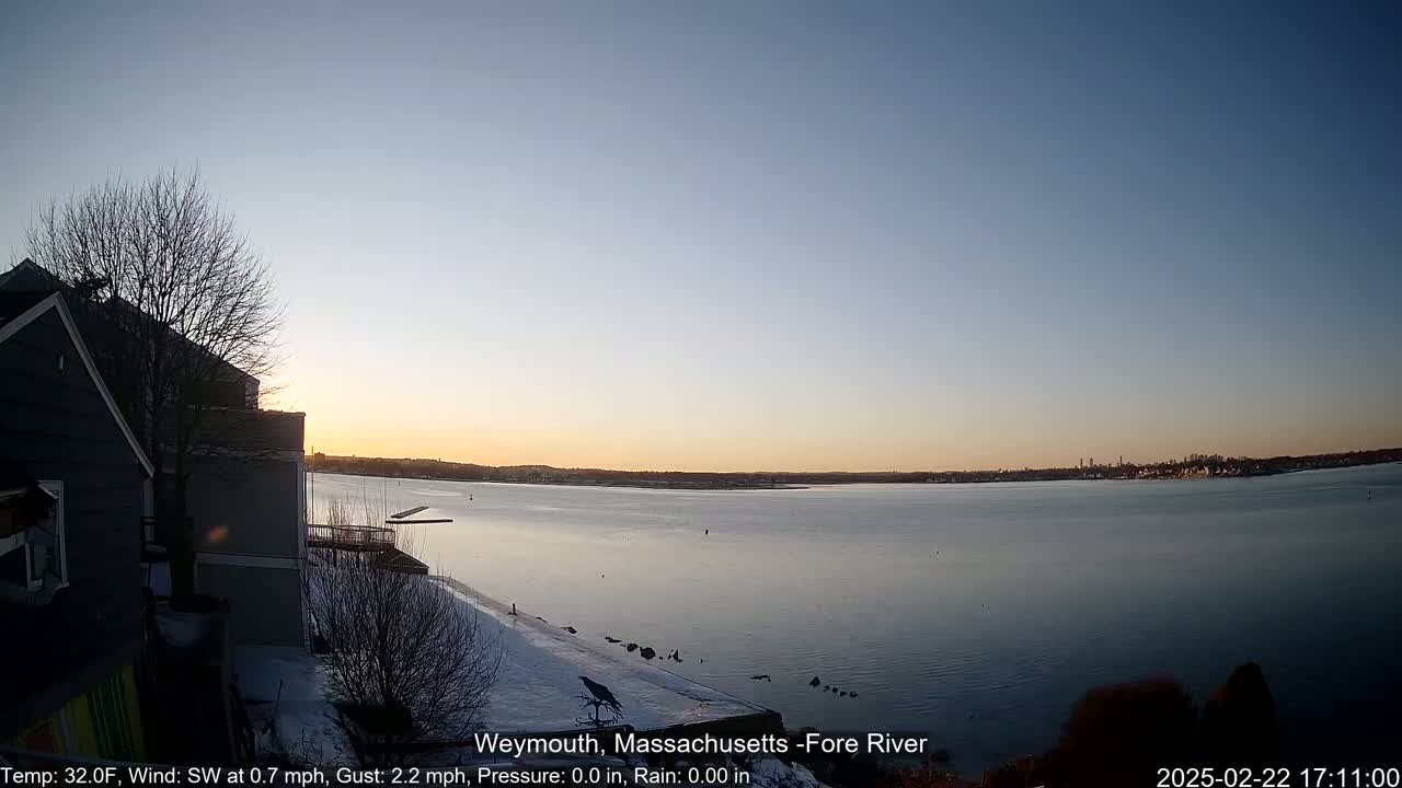 A calm, twilight view of a river from a snowy shoreline, with a house and bare trees in the foreground.
