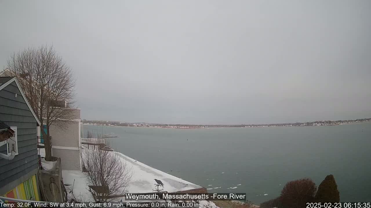 A snow-covered waterfront scene shows a calm body of water extending to a distant shoreline under an overcast sky.