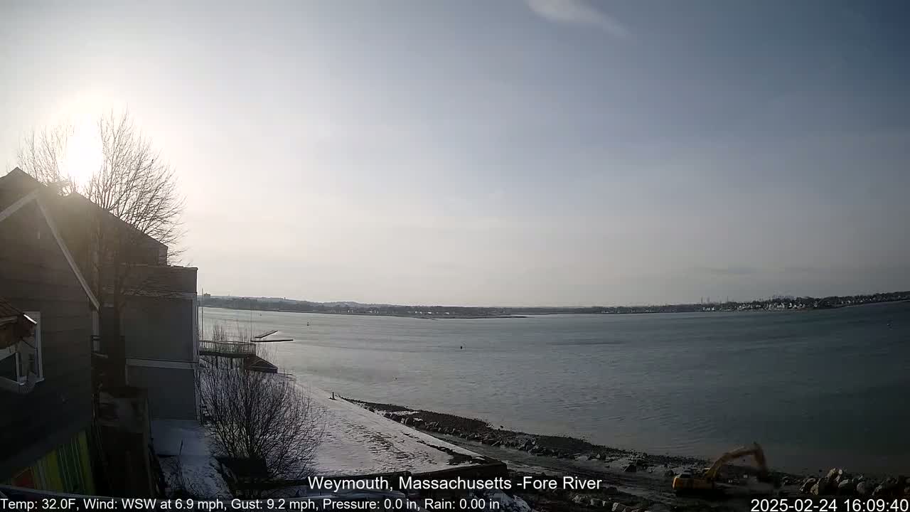 A sunny, cold day shows a calm body of water, a snowy shoreline, and a partially visible building in the foreground.