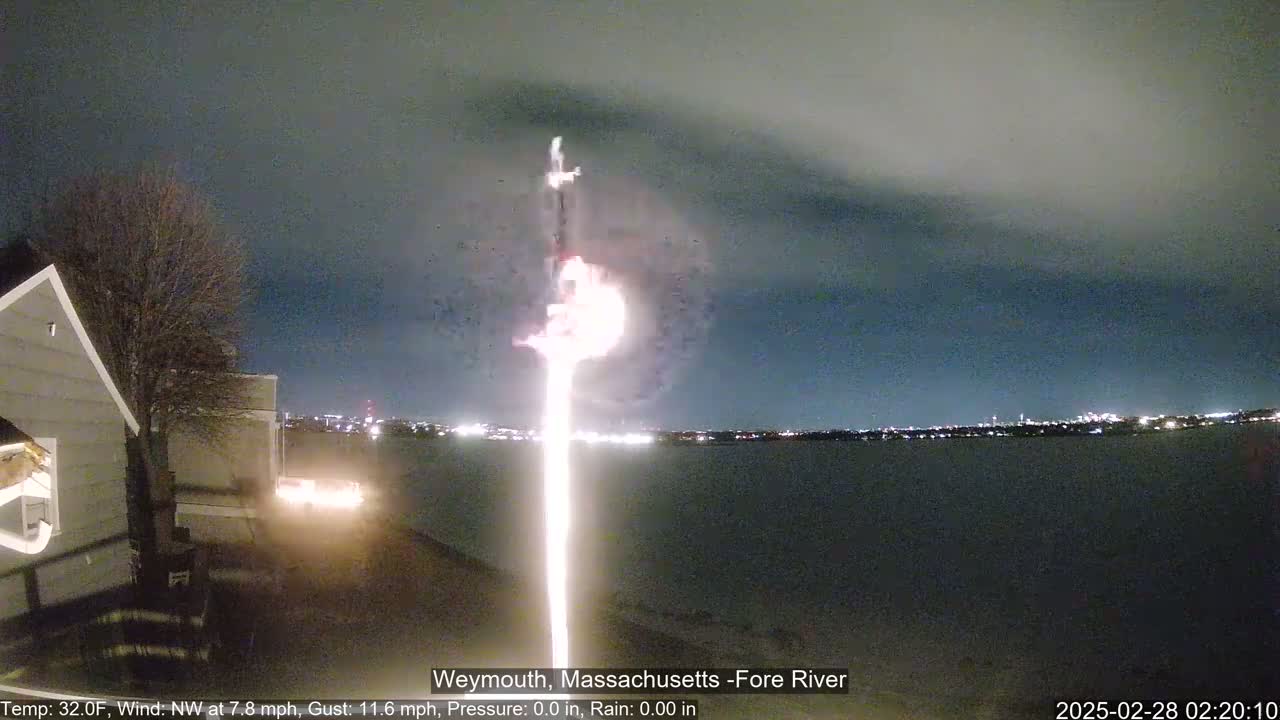 A bright, vertical light beam rises from a dark shoreline over a calm body of water under a nighttime sky.